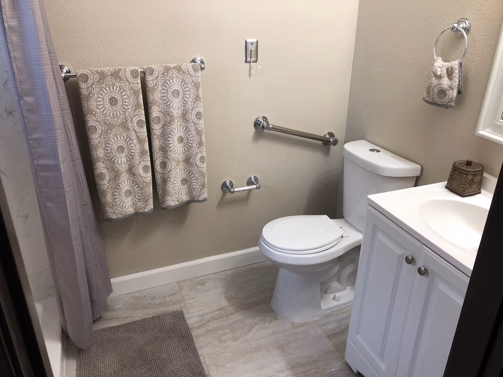 A small bathroom with a white toilet, white vanity with a sink, and a mirror above it. There are two beige towels with white floral patterns hanging on a towel rack next to the shower curtain. A matching hand towel hangs on a ring near the sink. The floor is tiled and there is a gray bath mat in front of the shower.