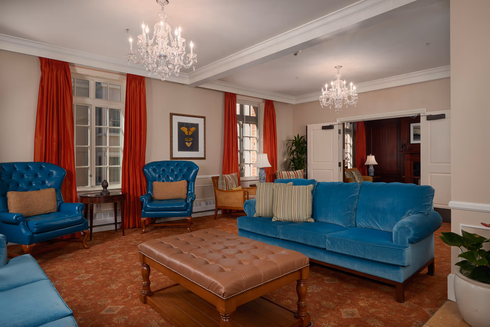 A cozy living room with blue upholstered sofas and armchairs, a large brown tufted ottoman in the center, red curtains on the windows, two chandeliers hanging from the ceiling, and a framed butterfly artwork on the wall.