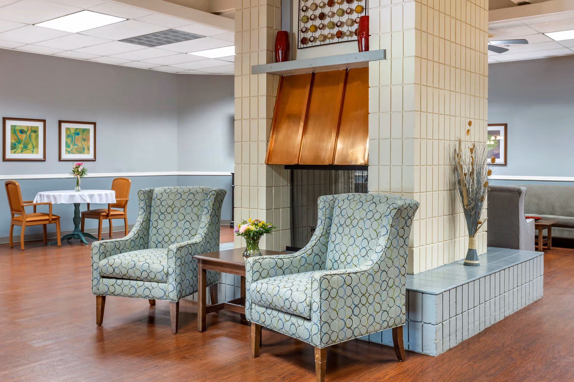 A cozy seating area in a senior living facility featuring two patterned armchairs with a small wooden table between them, holding a vase with flowers. The chairs are positioned in front of a tiled fireplace with a copper hood. In the background, there is a round table with two chairs and framed artwork on the walls. The room has wood flooring and soft lighting.