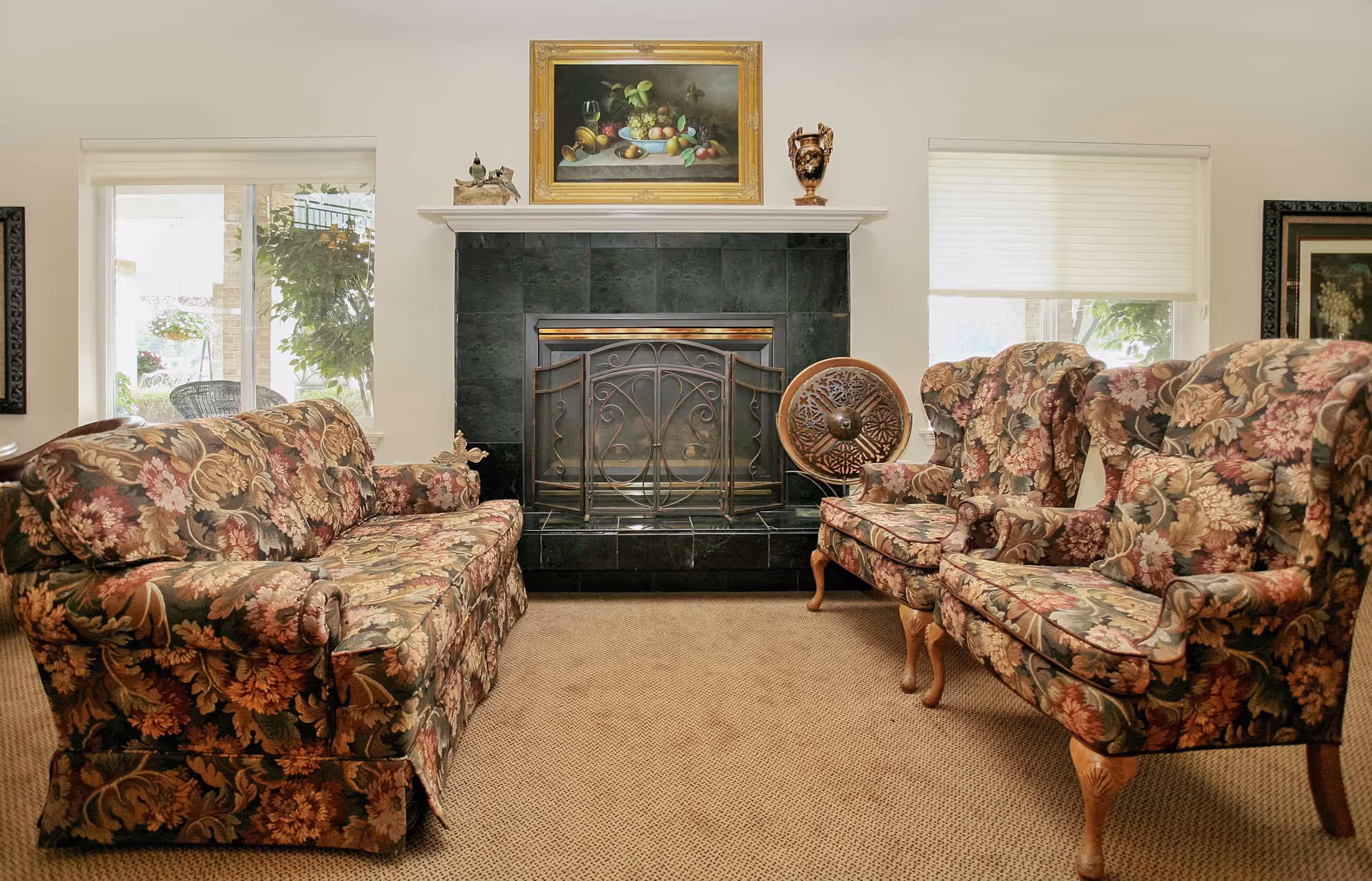 A cozy living room with floral patterned sofa and two matching armchairs facing a black tiled fireplace with a decorative metal screen. Above the fireplace is a framed painting of fruit and a decorative vase. Two windows with white blinds flank the fireplace, letting in natural light.