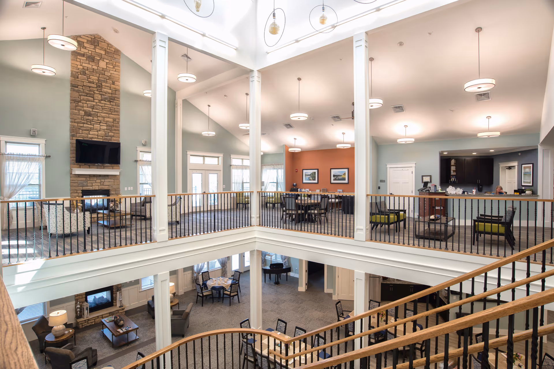 Interior view of a senior living facility showing a spacious two-story common area with seating arrangements, a stone fireplace with a mounted TV, multiple tables and chairs, large windows with sheer curtains, and a reception desk. The area is well-lit with modern ceiling lights and features a wooden staircase with black railings.