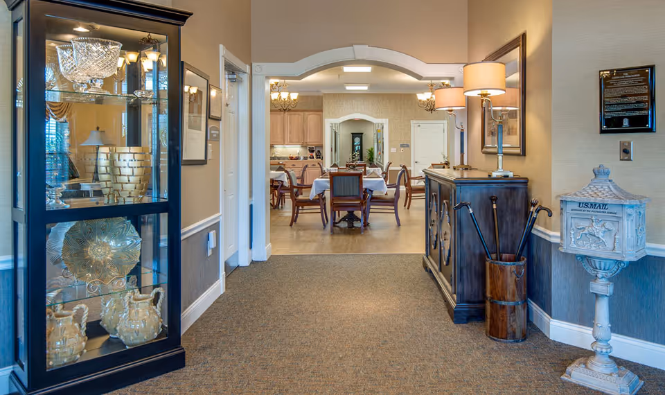 Entrance hallway in a senior living facility with a glass display cabinet on the left, a sideboard and lamp on the right, leading into a dining room with tables and chairs.