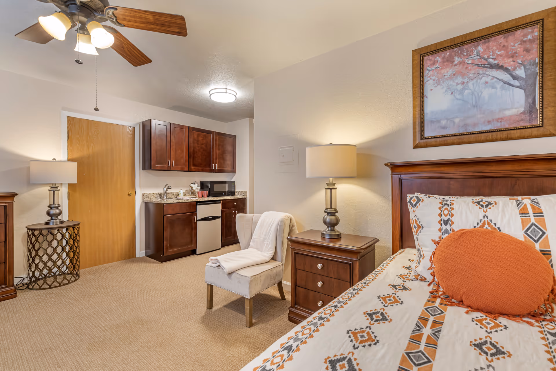 A cozy senior living facility room featuring a bed with patterned bedding and an orange round pillow, a wooden nightstand with a lamp, a cushioned chair with a white blanket draped over it, and a kitchenette area with dark wood cabinets, a small refrigerator, microwave, and sink. The room has beige carpet, a ceiling fan with lights, and a framed painting of a tree with red leaves on the wall.