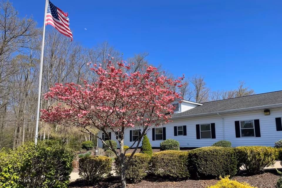 Exterior view of a white senior living facility building with black shutters, surrounded by neatly trimmed bushes and a blooming pink tree. An American flag is flying on a flagpole against a clear blue sky with some leafless trees in the background.