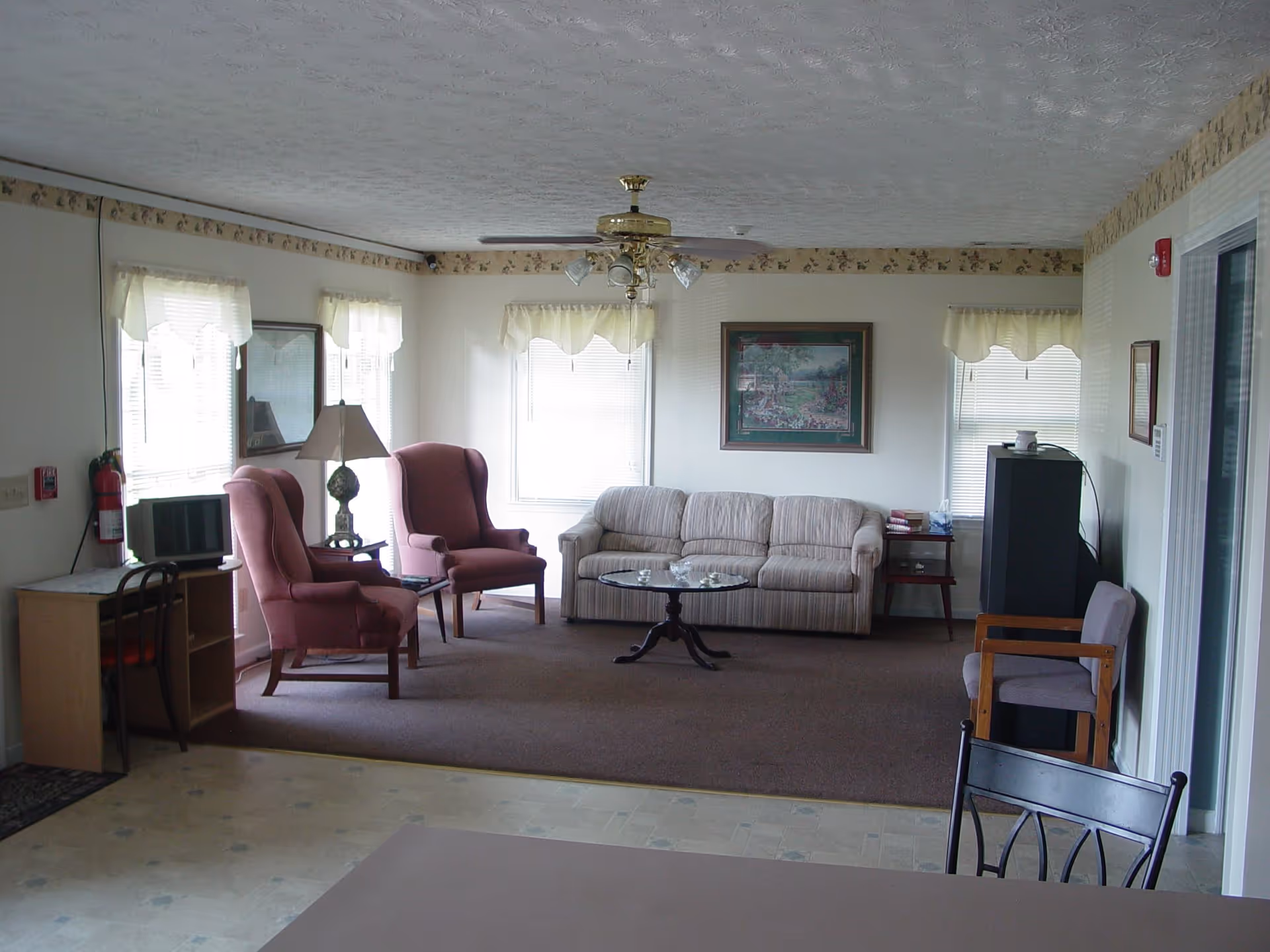 A cozy living room area with two pink upholstered armchairs, a beige striped sofa, a round glass coffee table, and a ceiling fan with lights. The room has several windows with white curtains, a framed painting on the wall, a small wooden desk with a chair and a microwave, a floor lamp, and a small side table with books and tissues.