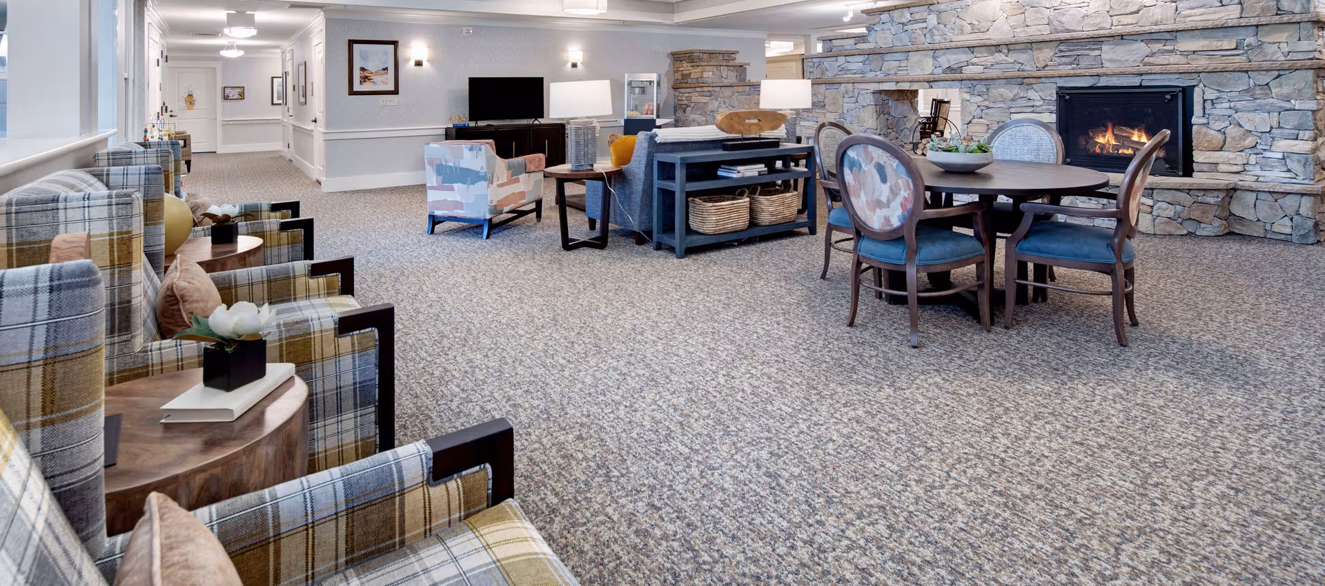 Communal lounge area with plaid armchairs, a round table, seating, TV console and a stone fireplace.