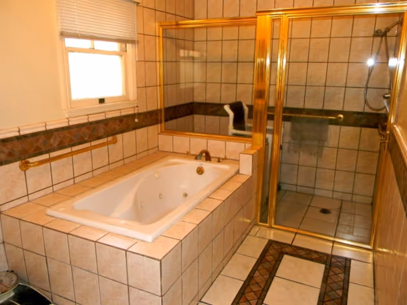 A tiled bathroom featuring a built-in bathtub with jets and a separate glass-enclosed shower with gold trim. The walls and floor are covered in beige tiles with decorative brown accents. There is a window with blinds above the bathtub and a grab bar on the wall beside the tub.