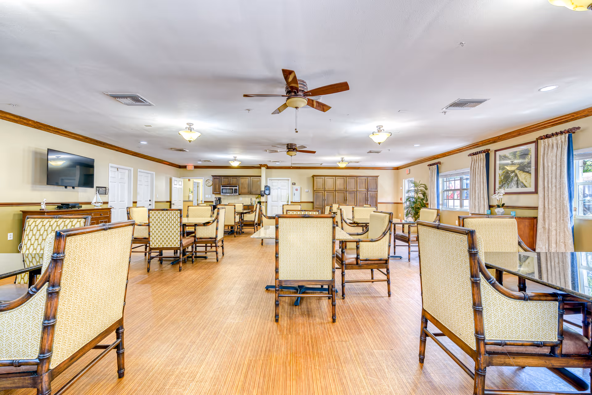 Large communal dining room with multiple tables and upholstered chairs, ceiling fans, a wall-mounted TV, and windows.