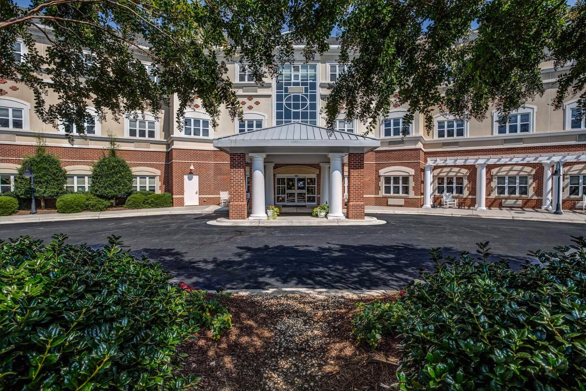 Front entrance of a senior living facility with a covered drop-off area supported by white columns. The building has a brick and beige facade with multiple windows. Green bushes and trees frame the entrance, and a paved driveway leads up to the door.
