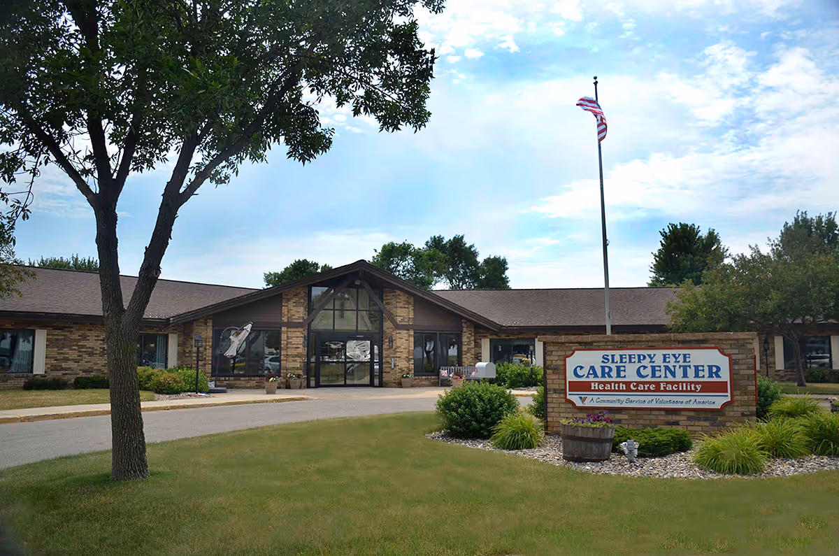 Exterior view of Sleepy Eye Care Center, a single-story brick building with large windows and a peaked roof entrance. There is a flagpole with an American flag in front of the building and a landscaped area with grass, bushes, and a sign that reads 'Sleepy Eye Care Center Health Care Facility'.