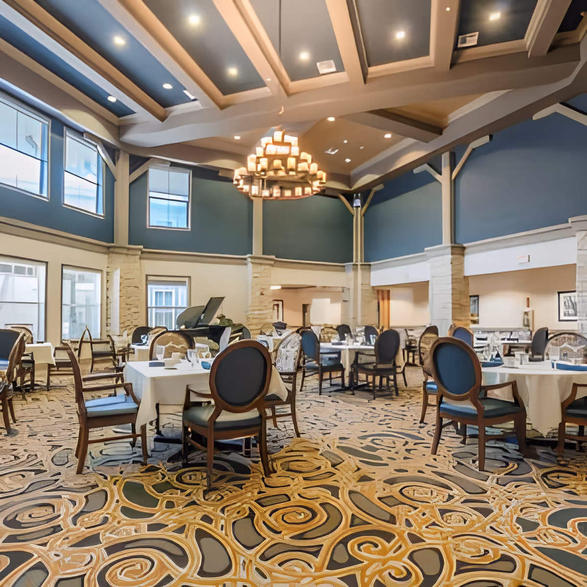 A spacious dining room with multiple tables covered with white tablecloths and set with glassware and napkins. The room features a high ceiling with exposed beams and a large chandelier in the center. The walls are painted blue with large windows allowing natural light to enter. The floor has an intricate patterned carpet, and there is a grand piano in the background.