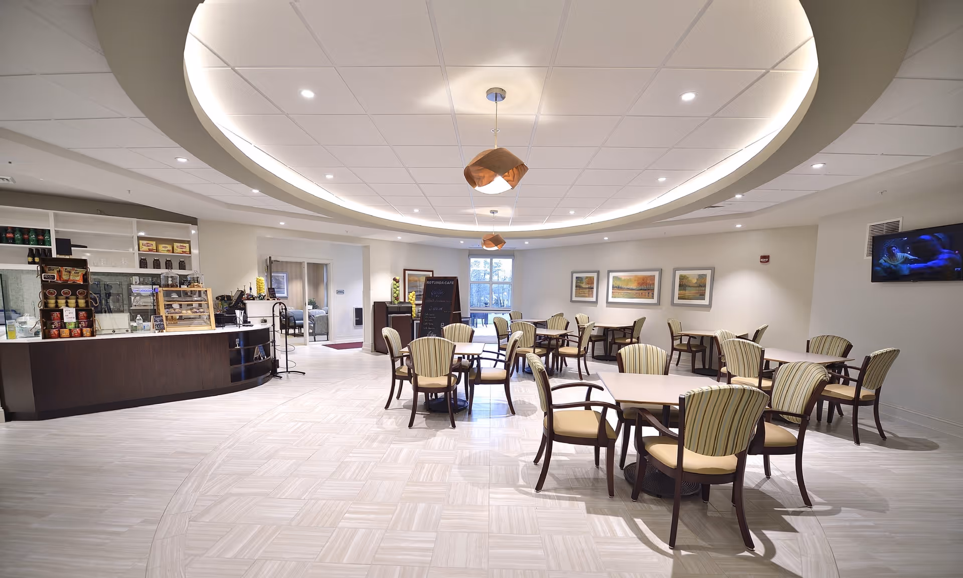 A spacious and well-lit dining area with multiple tables and chairs arranged neatly. The ceiling features a circular recessed design with modern pendant lights. On the left side, there is a counter area with shelves stocked with various items and a display case. The back wall has a window letting in natural light and framed artwork. A television is mounted on the right wall.