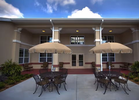 Outdoor patio area with two round tables, each surrounded by four chairs and shaded by large beige umbrellas. The patio is in front of a beige building with multiple windows and a double door entrance. There are plants on either side of the patio.