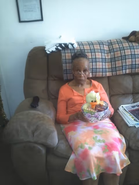 An elderly woman wearing glasses sits on a living-room couch holding a floral gift basket with a plush chick.