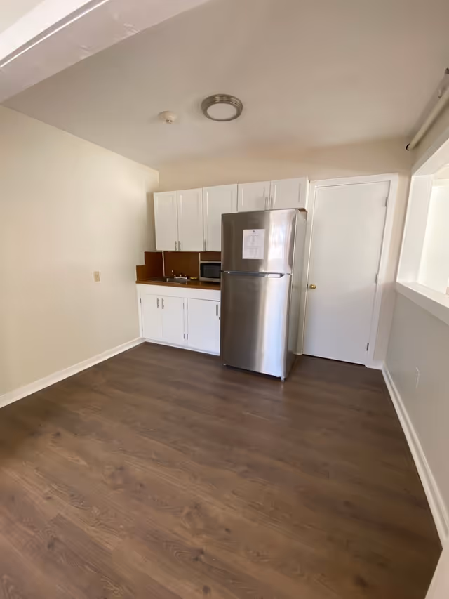 Small kitchenette with a stainless steel refrigerator, white cabinets, sink and microwave in a room with wood flooring.