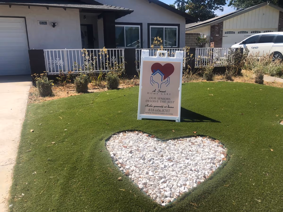 Front yard of a residential home with a green lawn featuring a heart-shaped area filled with white rocks. A white sandwich board sign stands on the lawn displaying the logo and contact information for A Sweet Home Care, a senior care service. The house has a white exterior with a small porch and a white railing, and a white vehicle is parked in the driveway.