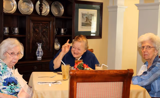 Three elderly women sitting around a dining table in a room with wooden furniture and decorative plates on a cabinet behind them. One woman is smiling and gesturing with her hand, while the other two look towards the camera. The table is set with glasses of iced tea and utensils.