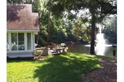 A peaceful outdoor scene at a senior living facility showing a green lawn, a large tree, a pond with a water fountain, and part of a building with large windows and a patio area with seating.