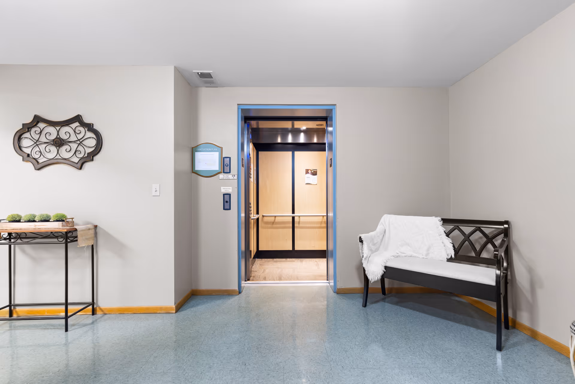 A clean and simple hallway area in an assisted living facility featuring an open elevator with wooden interior and metal handrails. To the right of the elevator is a black bench with a white cushion and a white throw blanket draped over it. To the left is a small decorative table with a tray holding small green plants, and a decorative wall hanging above it. The floor is covered with blue linoleum tiles and the walls are painted light beige.