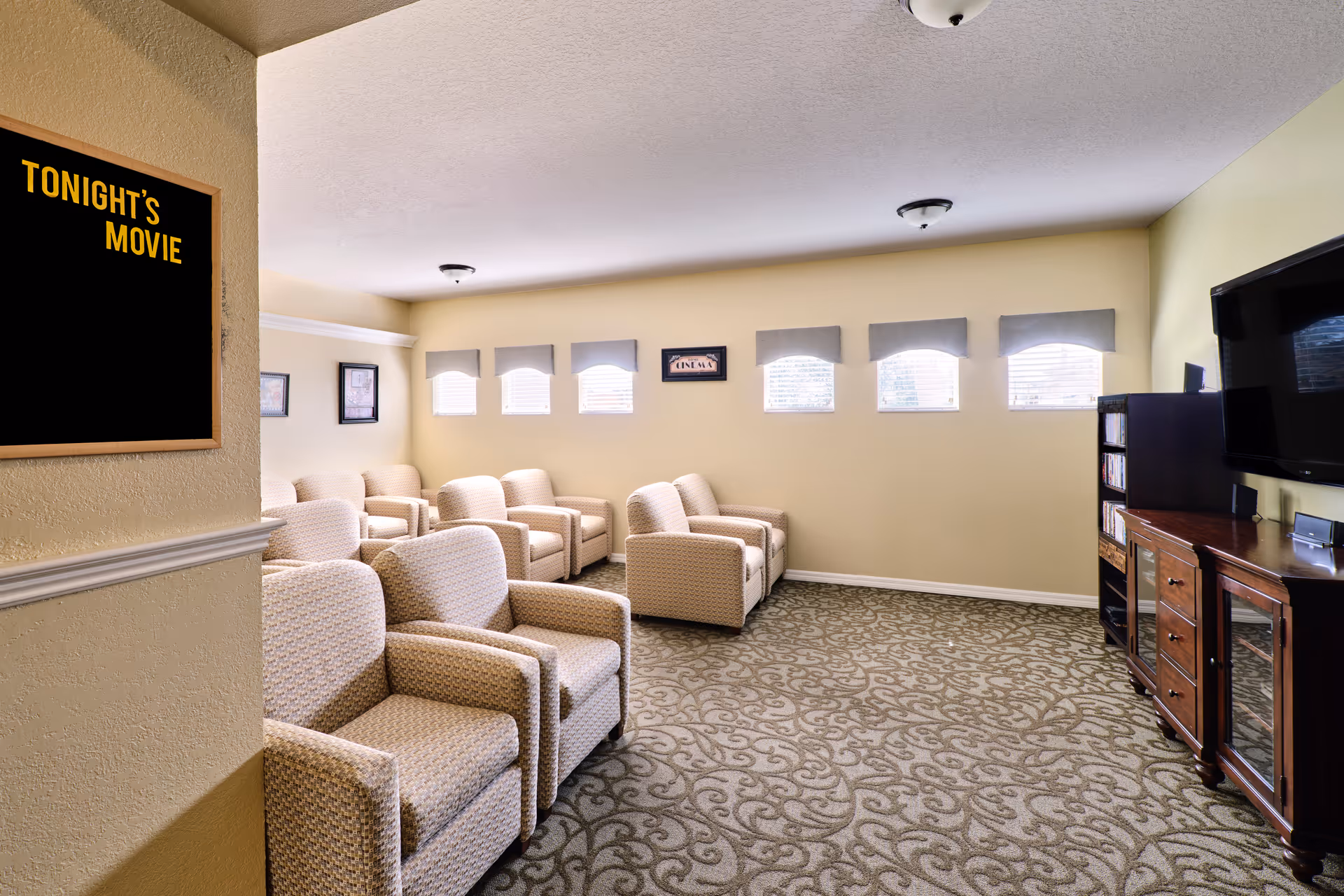 A cozy common room with multiple beige upholstered armchairs arranged in rows facing a flat-screen TV mounted on the wall. The room has a patterned carpet, several small windows with valances, and a sign on the wall that reads 'Tonight's Movie'.
