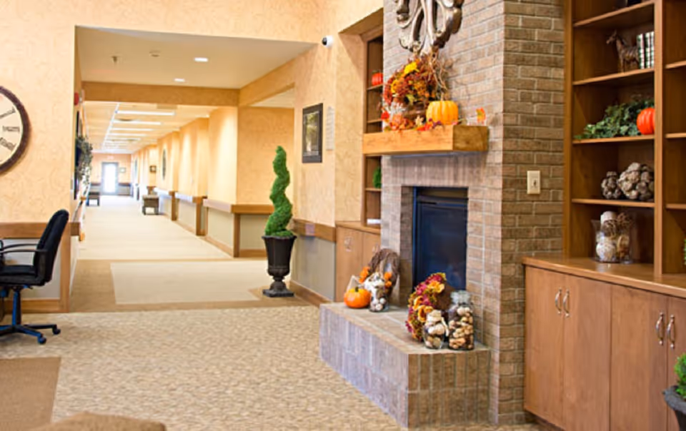A well-lit hallway in a senior living facility with beige walls and carpeted floors. On the right side, there is a brick fireplace decorated with autumn-themed items including pumpkins and fall leaves, surrounded by wooden shelves and cabinets with decorative plants and objects. A black office chair is visible on the left side near a large wall clock. The hallway extends into the distance with handrails along the walls.