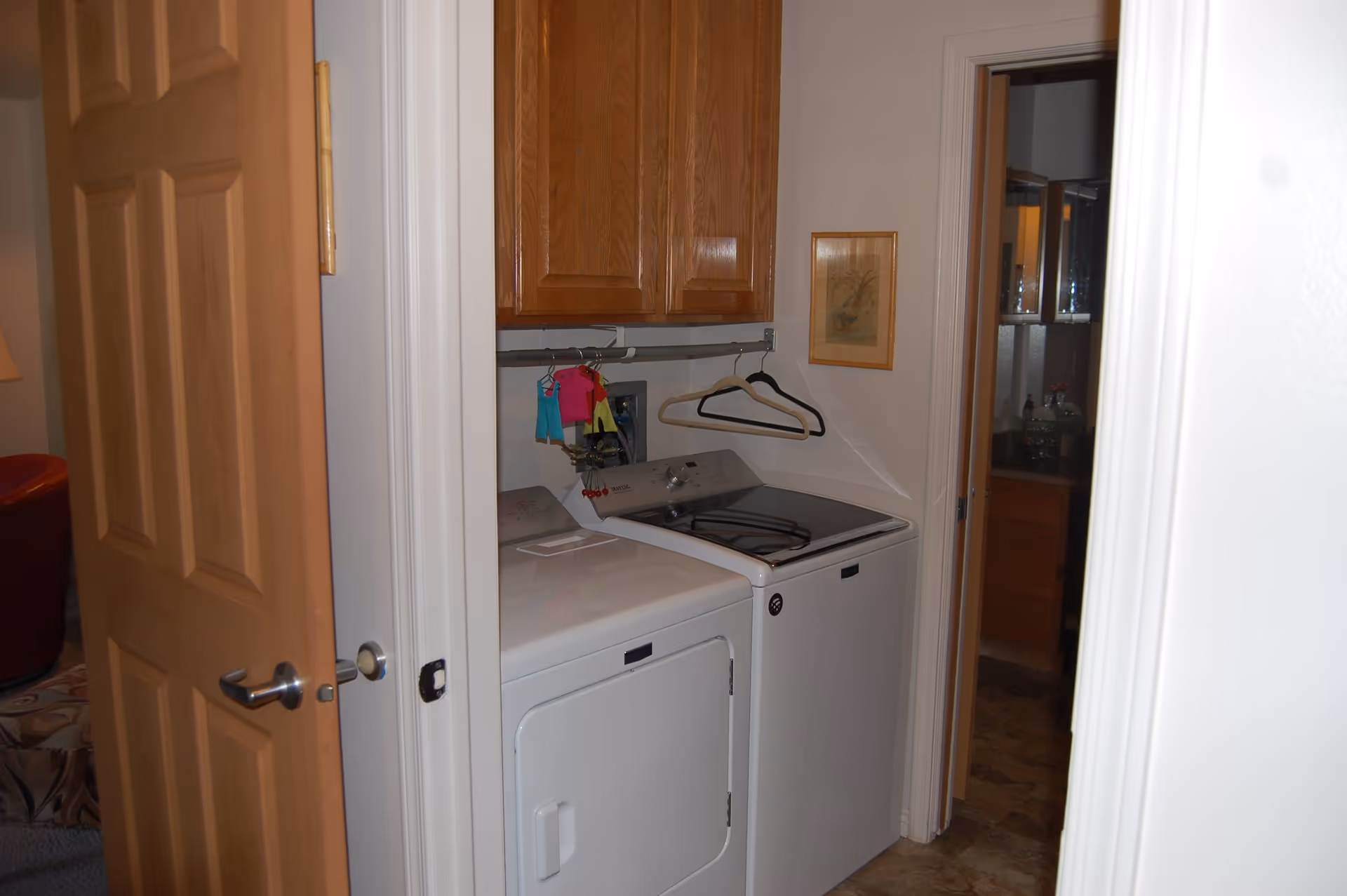 Small laundry area with a white washing machine and dryer side by side, wooden cabinets above, a few hangers and small clothes hanging on a rod, and a framed picture on the wall. The laundry area is adjacent to a bathroom visible through an open door.
