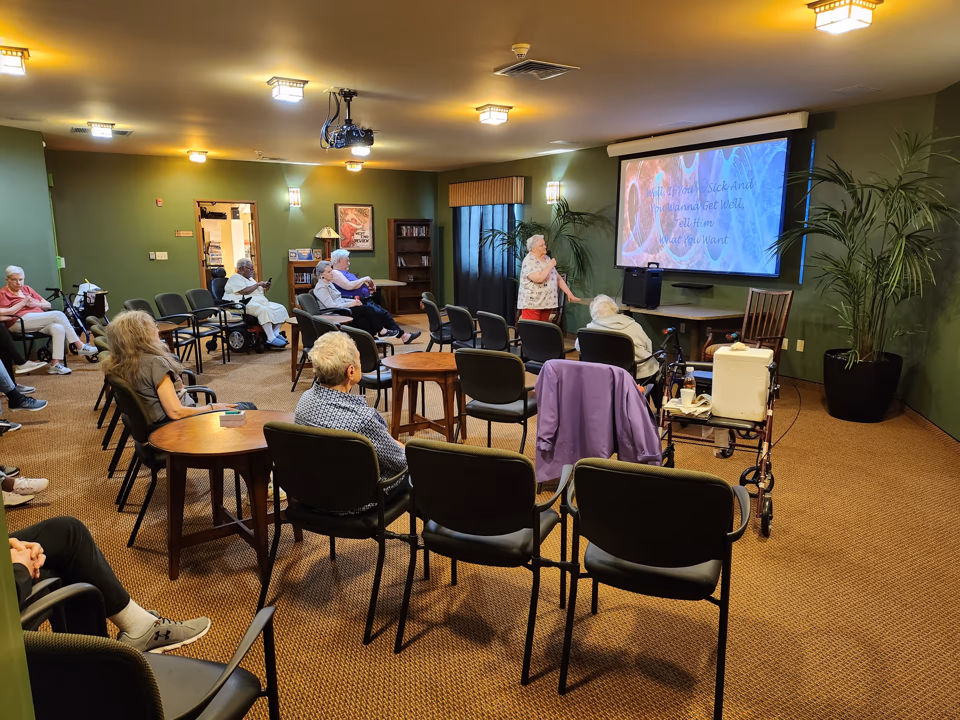 A group of elderly people seated in a room with green walls and brown carpet, watching a large screen at the front displaying song lyrics. One elderly woman is standing and singing into a microphone. The room has several chairs and tables, potted plants, and warm lighting.