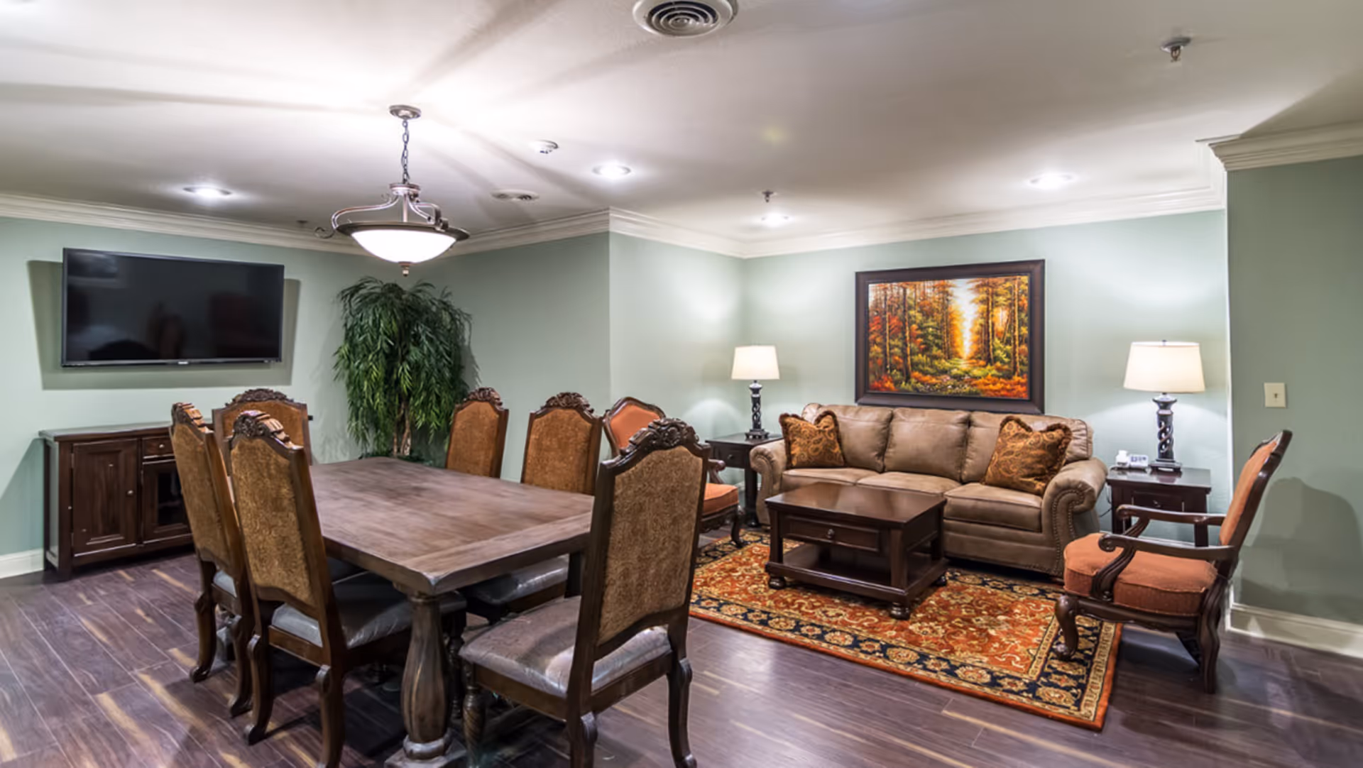 A furnished common room with a large dining table and chairs, a seating area with a sofa, armchairs, coffee table, rug, wall art, and a mounted TV.