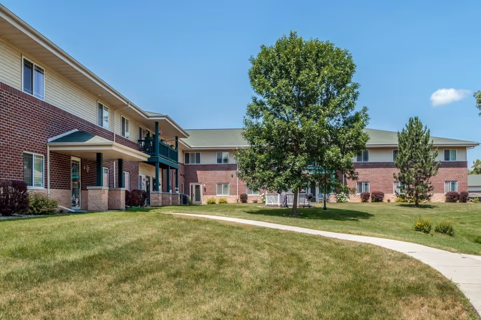 Exterior view of a two-story senior living facility building with red brick and beige siding. There is a green lawn with a concrete walkway curving through it, and several trees including a large leafy tree and a pine tree. The sky is clear and blue.