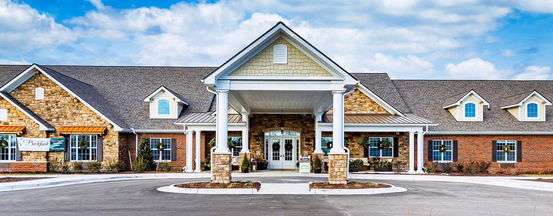 Front exterior view of Bickford of Canton facility with a large covered entrance supported by white columns, stone and brick facade, multiple windows with wreath decorations, and a clear blue sky with some clouds above.