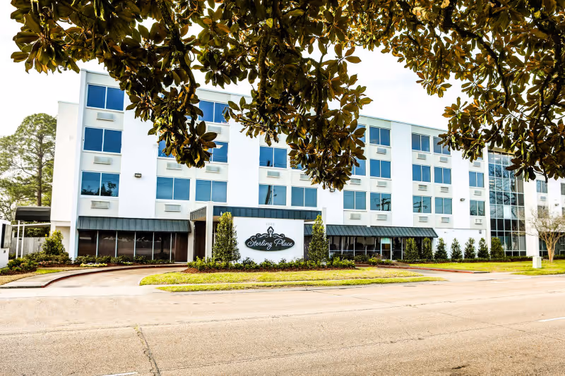 Front exterior of a four-story white senior living building with a 'Sterling Place' sign and tree branches in the foreground.