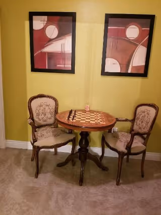 A small round wooden table with a built-in chessboard on top, flanked by two upholstered vintage-style chairs with carved wooden frames. The table and chairs are set against a yellow wall with two abstract red and white paintings hanging above them. The floor is tiled in a light color.