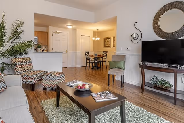 A cozy living room area with a beige sofa, a patterned armchair with matching ottoman, a dark wooden coffee table with magazines and a bowl of fruit, a green rug, a flat-screen TV on a wooden stand, a decorative round mirror on the wall, and a small dining area with a round table and four chairs in the background.
