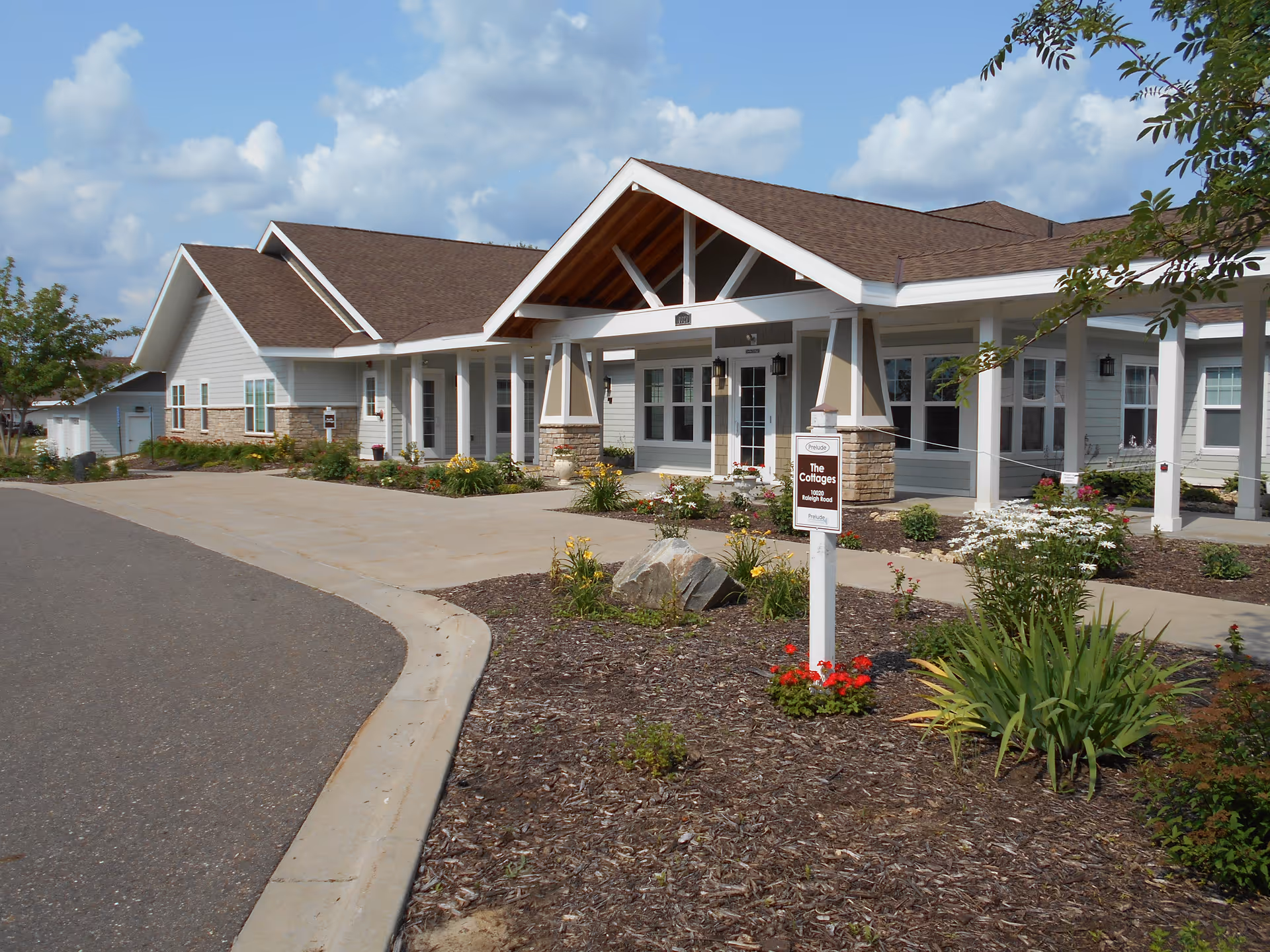 Exterior view of a single-story building with a brown roof and white siding, featuring a covered entrance supported by columns. There is a paved driveway and landscaped garden beds with flowers and shrubs in front of the building under a partly cloudy sky.