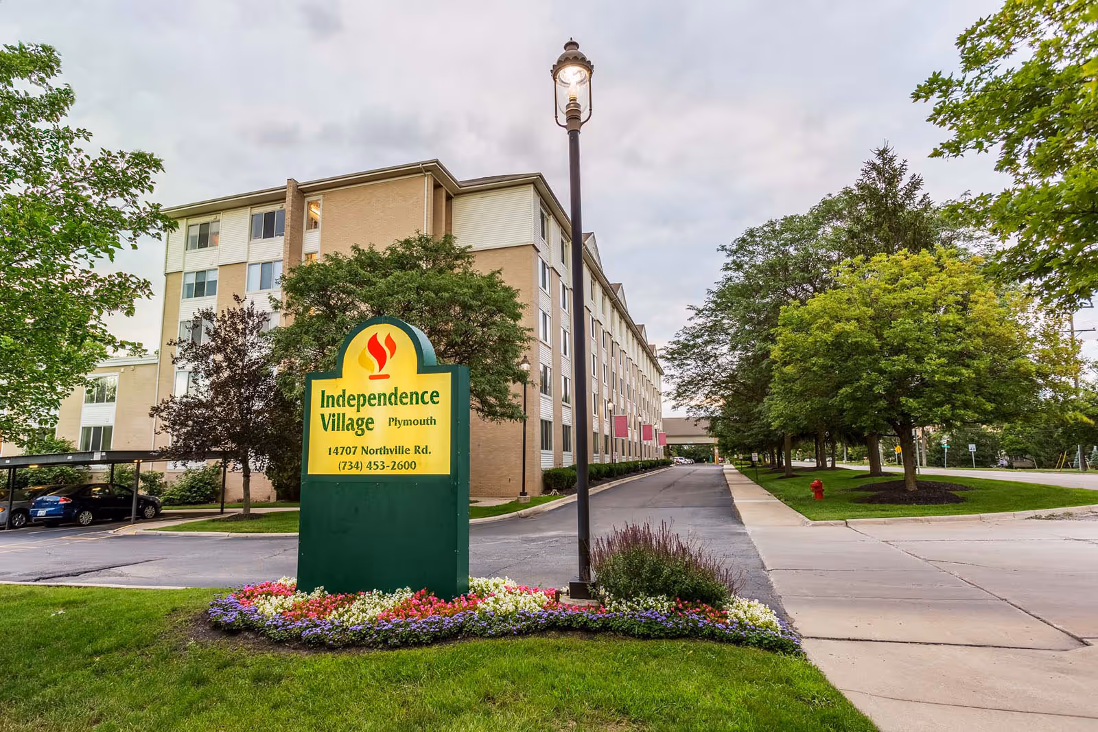 Exterior view of Independence Village of Plymouth, showing a multi-story building with a driveway and a landscaped area with colorful flowers surrounding a green and yellow sign displaying the facility's name and contact information.