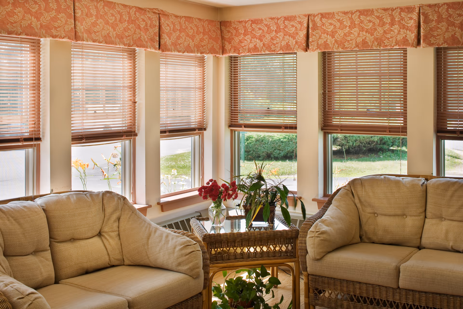 A cozy sitting area with two beige cushioned wicker sofas facing each other, separated by a glass-top wicker table with a vase of red flowers and a potted plant. The room has multiple windows with wooden blinds and floral valances, allowing natural light to brighten the space.