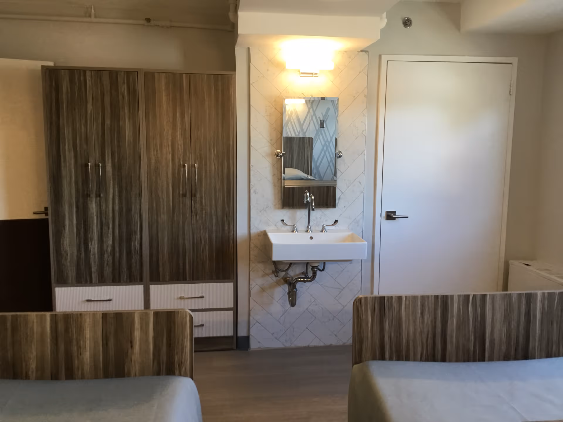 Interior view of a room with two beds featuring wooden headboards, a wall-mounted sink with a mirror above it, a wooden wardrobe with drawers, and a closed white door.