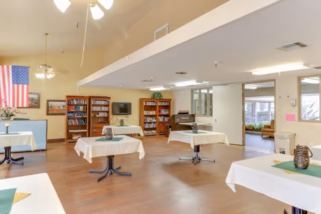 Spacious communal dining/activity room with several small tables covered in white tablecloths, bookshelves, a TV, piano, and an American flag.