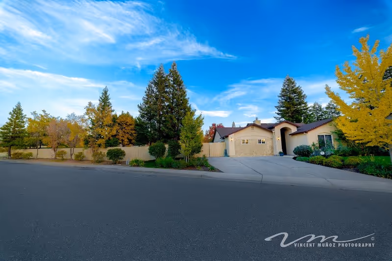 Exterior view of a single-story residential building with a driveway and garage, surrounded by trees with autumn foliage under a blue sky with scattered clouds.