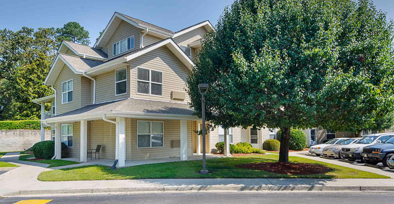 Exterior view of a two-story beige residential building with white trim, surrounded by green grass, a large leafy tree, and a parking area with several cars parked. The sky is clear and blue.