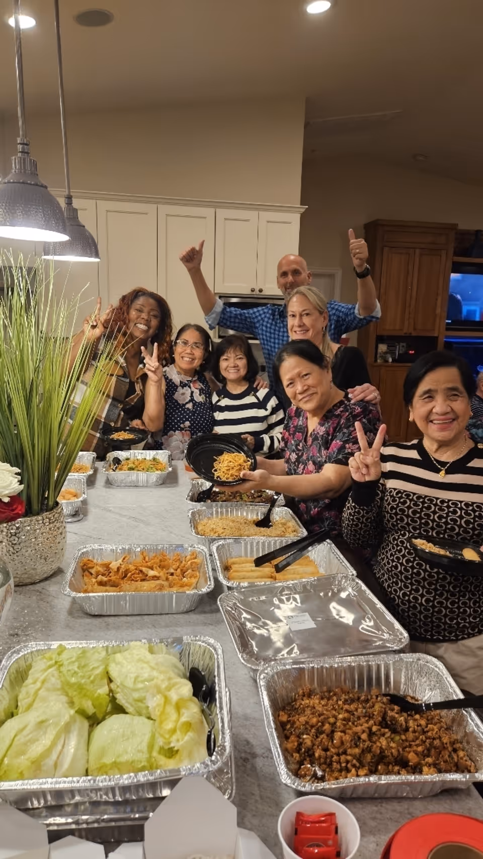 A group of seven smiling adults gathered around a kitchen island with various trays of food, including lettuce, noodles, and other dishes. They are posing for the photo with peace signs and thumbs up gestures. The kitchen has white cabinets and pendant lights hanging above the island.