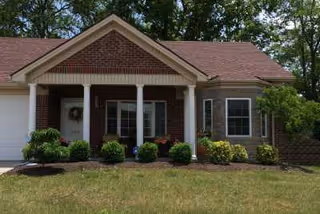 Front exterior view of a single-story brick house with a covered porch supported by white columns, surrounded by green shrubs and trees.