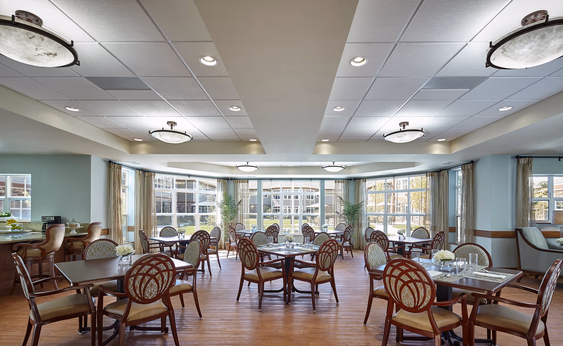 A bright and spacious dining room with multiple wooden tables and chairs arranged neatly. Large windows allow natural light to fill the room, and there are plants near the windows. The ceiling has recessed lighting and decorative light fixtures. The floor is wooden, and there is a small bar area with high chairs on the left side.