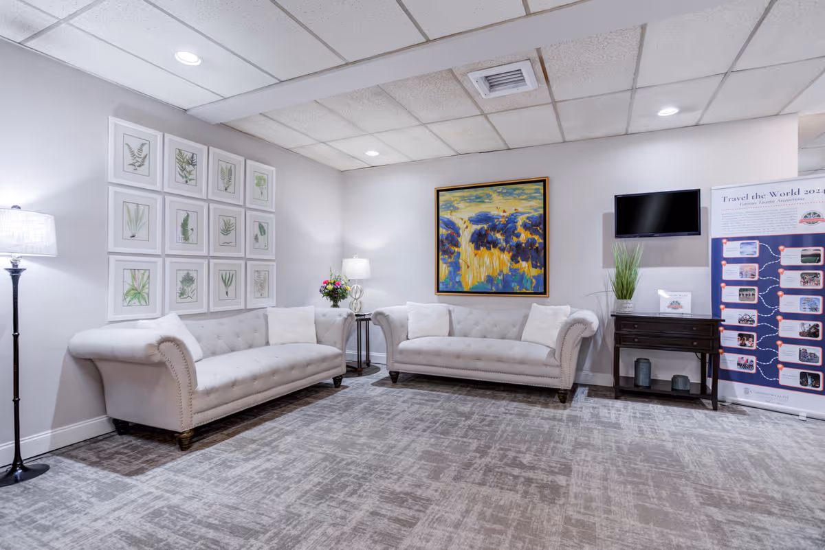 Bright lounge area with two tufted sofas, framed artwork, a wall-mounted TV, and side tables with lamps and plants.