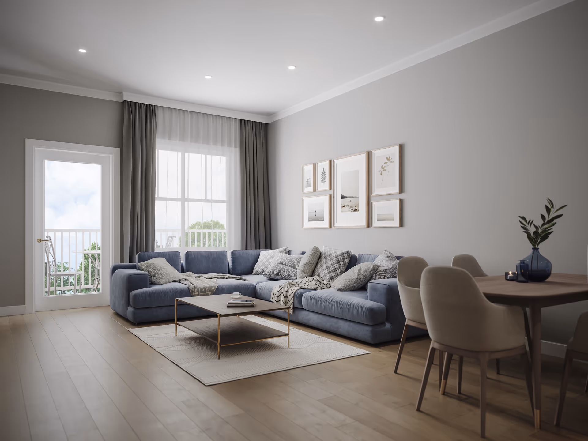 A modern living room with a large blue sectional sofa adorned with multiple patterned cushions and throws. A rectangular coffee table sits on a light-colored rug in front of the sofa. On the right side, there is a wooden dining table with four beige upholstered chairs and a blue vase with green leaves as a centerpiece. The room has light wooden flooring, gray walls, and a large window with sheer white curtains and darker drapes. A glass door leads to an outdoor balcony with white railings and a small table with chairs. Several framed pictures hang on the wall above the sofa.