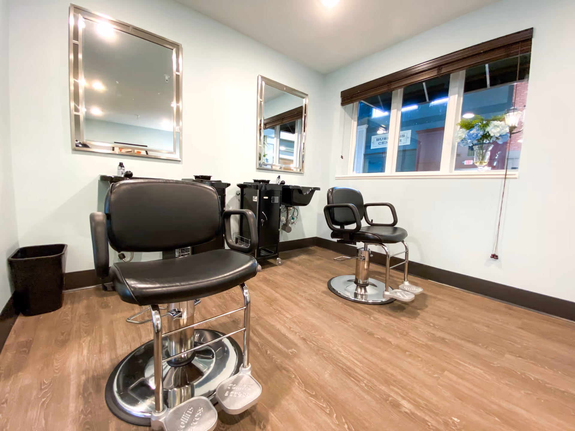 Interior salon room with two black barber chairs, mirrors, shampoo sinks and a window with flowers.
