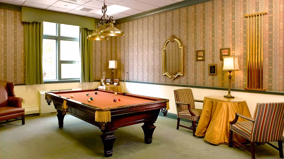 A cozy game room with a billiards table in the center, several billiard balls on the table, a hanging light fixture above it, a window with green curtains, a red armchair, a round table covered with a yellow cloth, two striped armchairs, a decorative mirror on the wall, and a rack holding pool cues.