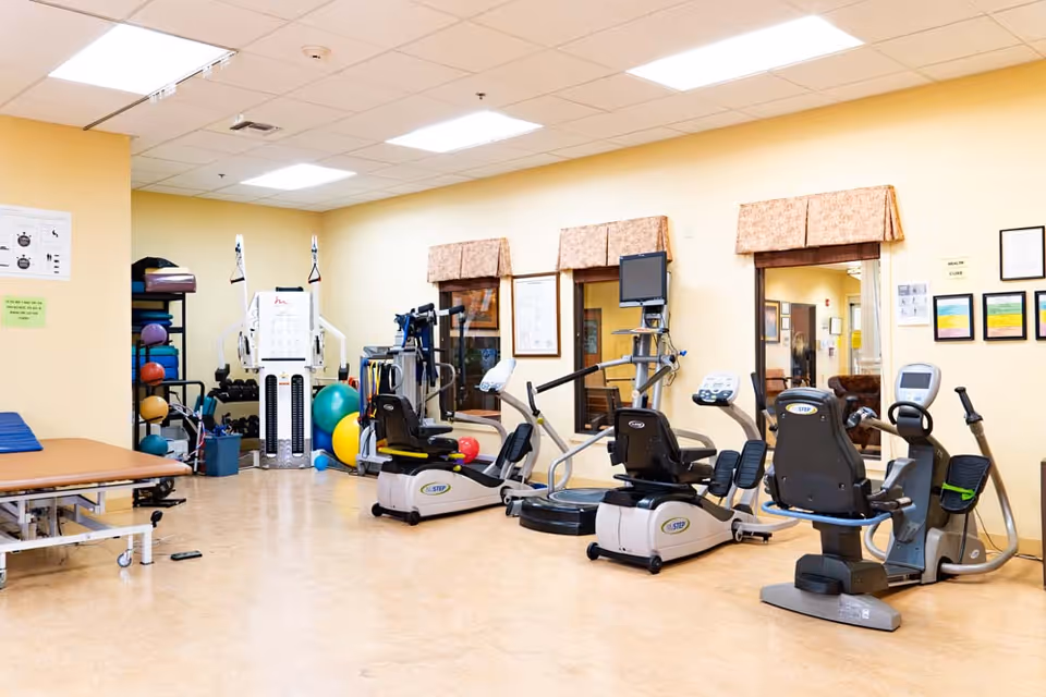 A rehabilitation exercise room with various fitness equipment including recumbent bikes, a multi-station weight machine, exercise balls, and dumbbells. The room has beige walls, a light-colored floor, and windows with valances. There are framed certificates and informational posters on the walls.