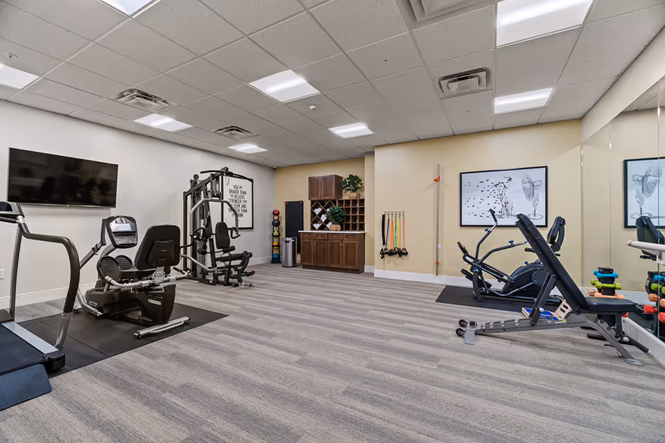 A senior living facility exercise room with various fitness equipment including a treadmill, recumbent bike, weight machine, and elliptical. The room has light-colored walls, a large mirror on one side, a wall-mounted TV, and a cabinet with plants and exercise accessories like resistance bands and dumbbells.