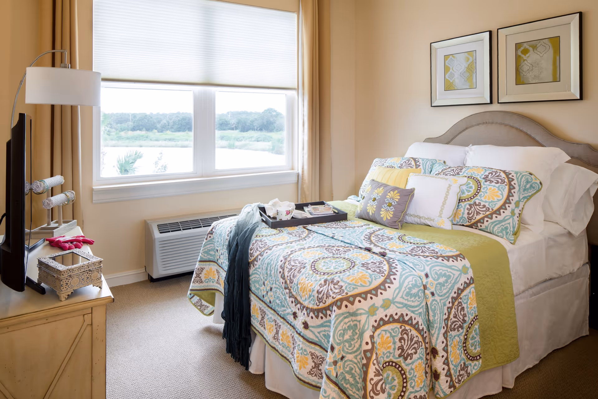 A bright and cozy bedroom with a neatly made bed featuring a colorful patterned quilt and multiple pillows. A tray with a cup and some items is placed on the bed. The room has a large window with a view of greenery outside, beige walls, and two framed artworks above the headboard. A TV is positioned on a wooden dresser with a decorative box and a lamp beside it.